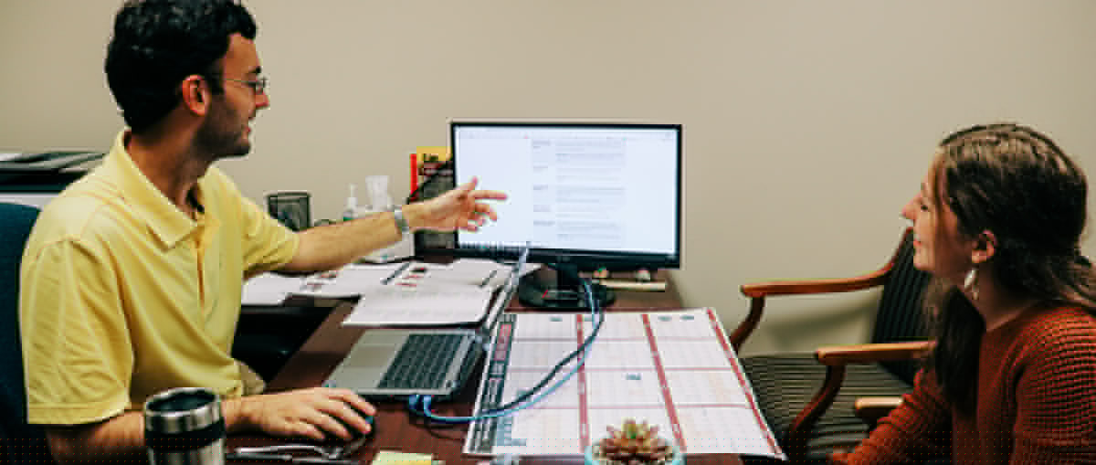 An advisor sits at a desk with a student, pointing to information on a computer screen