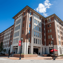 Students walking and on bikes walk on a brick courtyard. Behind them is a six story brick residence hall flanked by trees.