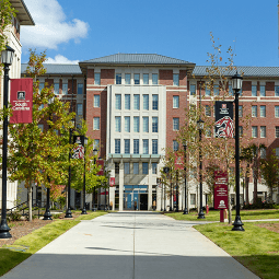 Students walking and on bikes walk on a brick courtyard. Behind them is a six story brick residence hall flanked by trees.