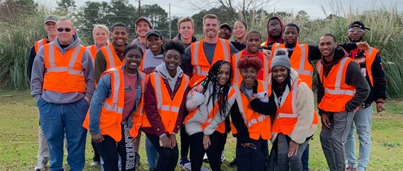 A diverse group of service volunteers wearing fluorescent vests pose in front of a newly cleaned patch of grass.