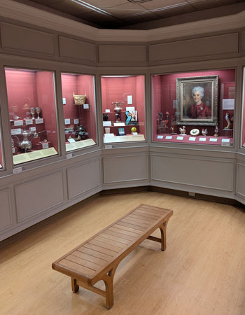 Small museum exhibit room with wood floors and a wooden bench in the center; glass display cases line the walls, showcasing historical artifacts and a portrait painting.