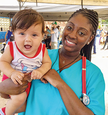 Shir’Mel McCullough wearing blue scrubs and a red stethoscope while holding a baby and smiling at the camera.
