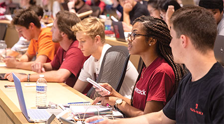 A row of students in class taking notes and looking to the front of the room.
