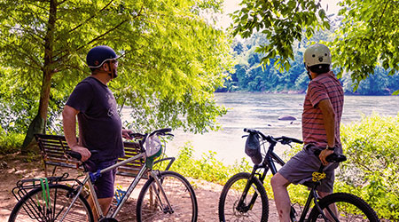Two people with bikes stopped on a wooded trail overlooking the river.