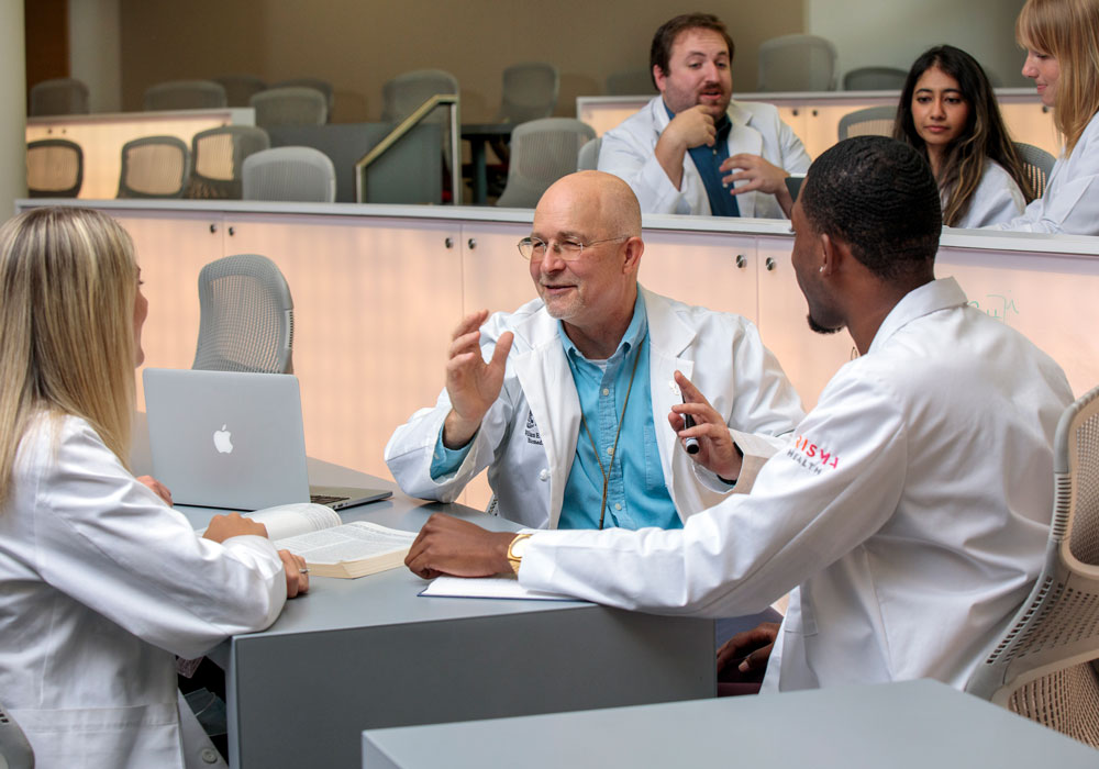 A professor speaks to a group of students seated a table in a large classroom.