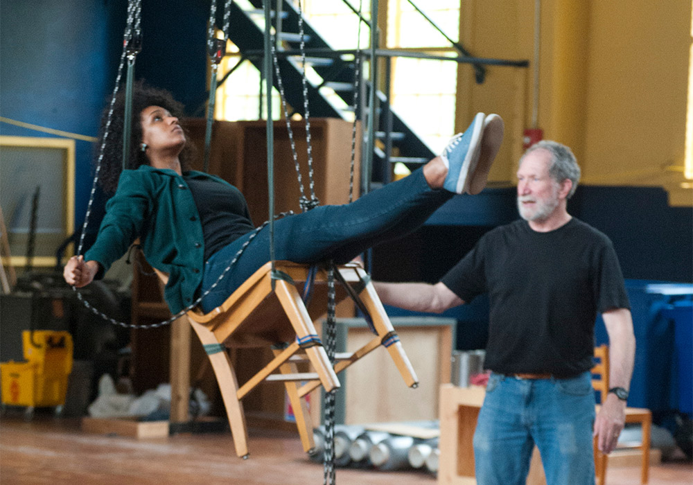 Student sits in a chair suspended in the air with ropes behind the scenes of a theatre production.