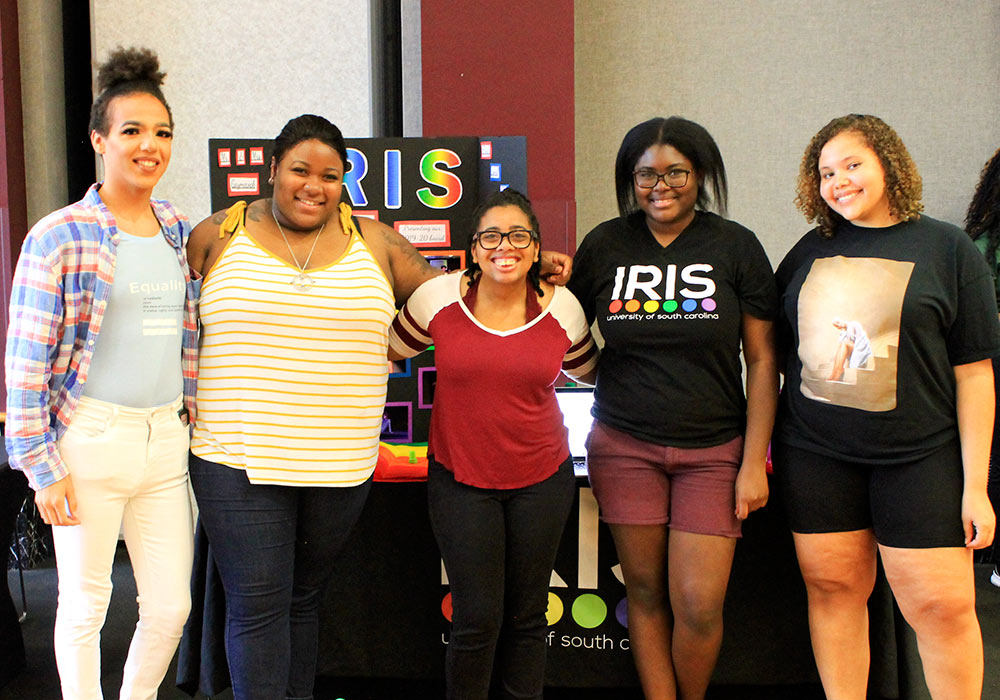Four students standing in front of an IRIS display board at a student organization fair.