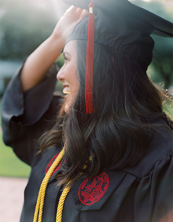 Student wearing graduation cap and gown adjusting her hat smiling in the sun.