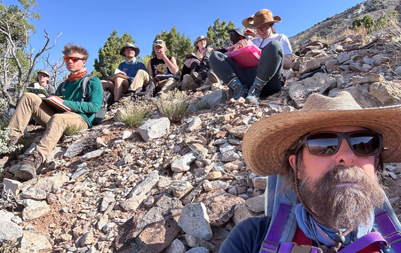 Geology professor David Barbeau takes a selfie while leading a group of University of South Carolina students sitting on a rocky hillside during field camp, with notebooks in hand and mountains in the background.