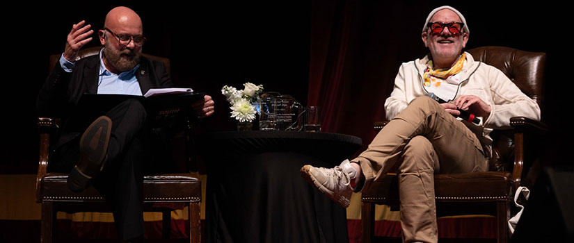 Michael Stipe, seated on the right in a white hoodie, scarf, and red sunglasses, smiles during a conversation on stage with a bald man in a dark suit holding a folder. They are seated in armchairs on either side of a small round table with flowers, water glasses, and an award plaque.