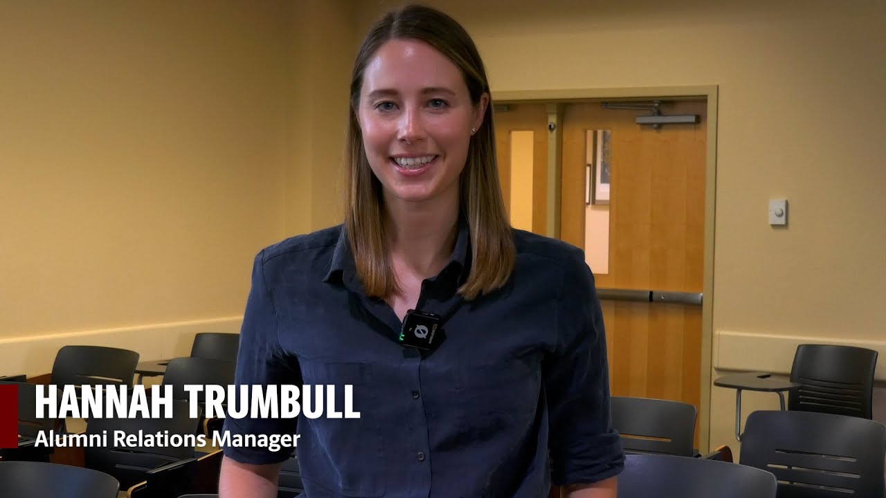 Hannah Trumbull, Alumni Relations Manager, smiles while standing in a classroom with rows of empty black chairs and double wooden doors in the background.