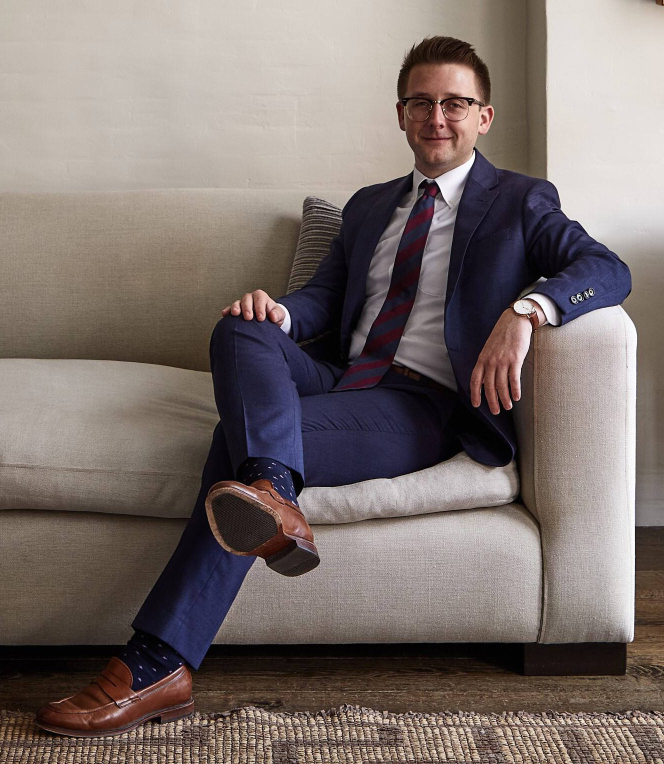 Aaron Anderson, wearing a navy suit and tie, sits confidently on a light-colored couch beneath a framed painting in a refined, art-filled interior