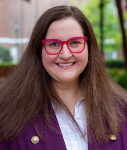 A photo of Alison Houge standing outside smiling with a purple jacket on and red glasses.