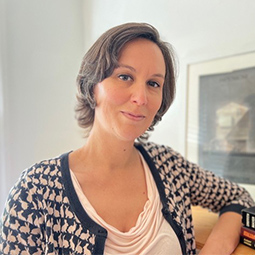 Woman with short brown hair wearing a patterned cardigan and light pink top, standing indoors next to a bookshelf and framed artwork.