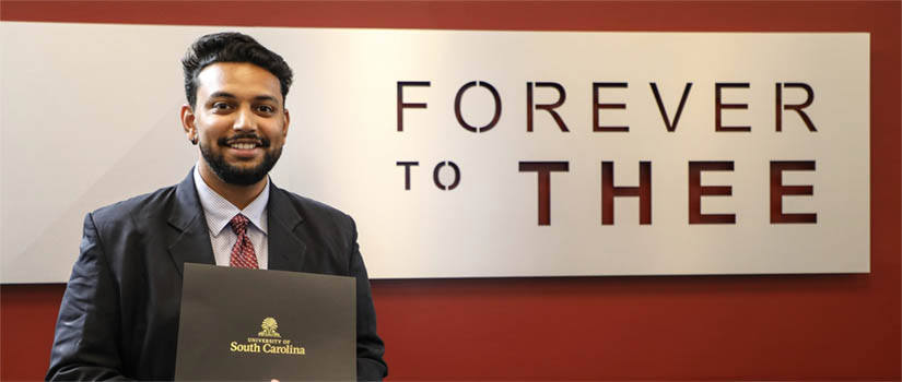smiling student holding award in front of a sign that reads "Forever to Thee"