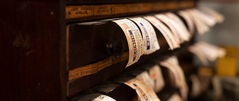 Close-up of an antique wooden drawer cabinet with labeled drawers, some pulled open to reveal vintage paper slips or tags hanging out.