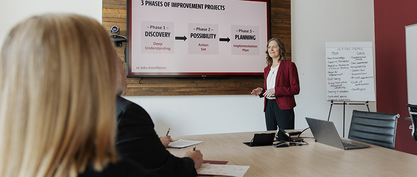 A woman in a red blazer gives a presentation to colleagues seated at a conference table, with slides and a flip chart of action items visible.