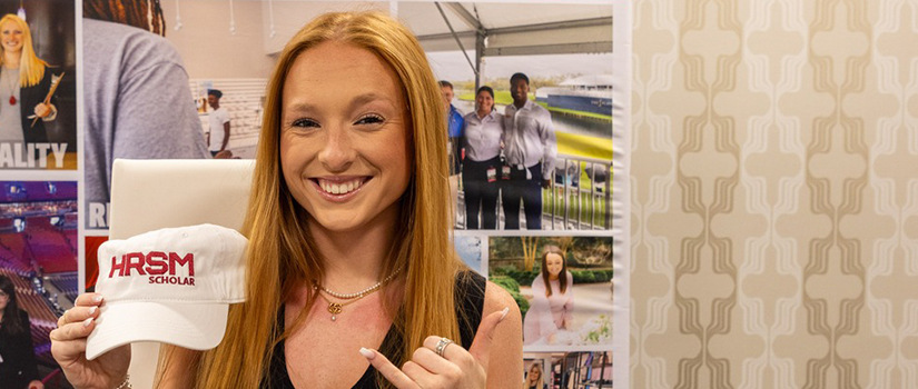 A smiling young woman with long red hair holds up a white hat that reads “HRSM Scholar.” She gestures with a Spurs Up sign and stands in front of a collage of photos showcasing hospitality, retail and sport management scenes.
