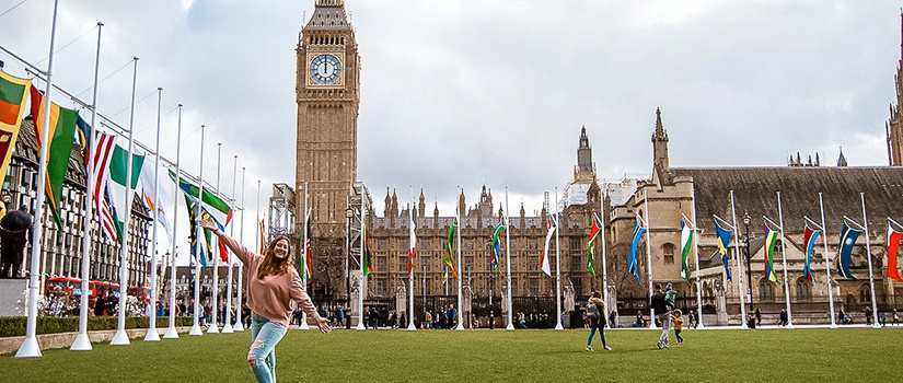 A young woman poses and smiles in front of the Palace of Westminster and Big Ben in London, surrounded by rows of colorful international flags on a grassy square.