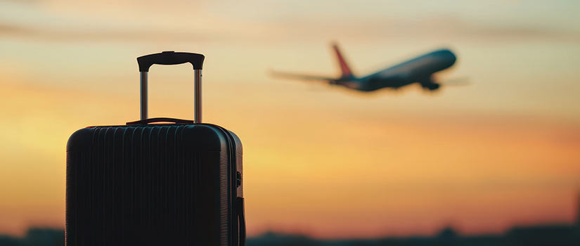 Black suitcase in the foreground with a commercial airplane taking off at sunset in the background.