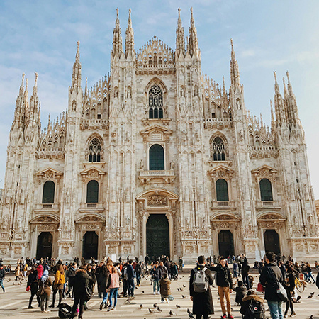 Crowds of people gather in front of the ornate Gothic facade of Milan Cathedral (Duomo di Milano) in Italy, with pigeons scattered across the square under a clear blue sky.