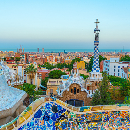 A view from Park Güell in Barcelona showcasing Gaudí’s colorful mosaic designs in the foreground and the city skyline stretching to the sea.