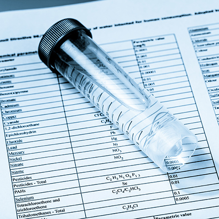 Clear sample vial filled with water resting on a printed chemical analysis report listing contaminants and measurement values.