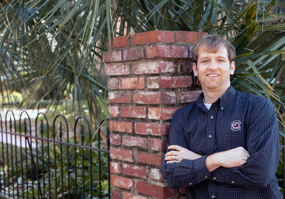 man leans against a brick wall with greenery in the background