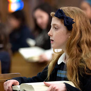 A red-headed girl in Catholic school uniform pays attention in class.