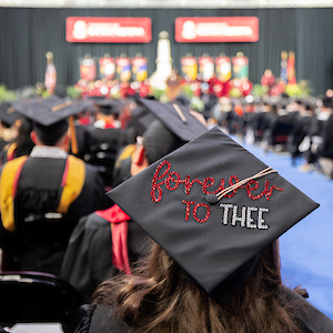 woman sitting in crowd wearing black graduation cap with glittery lettering that reads 